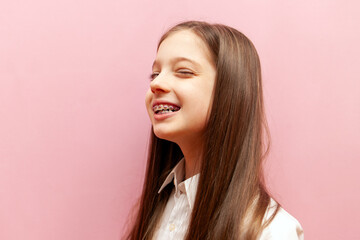 cheerful teenage girl with braces winks and hints on pink isolated background, happy child blinks...