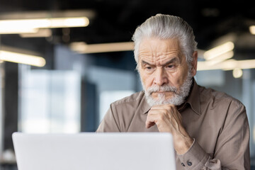 A senior man is deeply focused while using a laptop in a modern office, showing concentration and professionalism