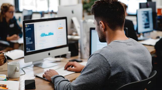 A young professional analyzes data on a computer screen in an open office environment. Colleagues are visible working in the background, creating a collaborative workspace.