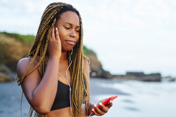Young woman with dark skin listening music in earhones connected to smartphone standing on beach washed by ocean and preparing for sport training.Afro american jogger in active wear and phone in hand
