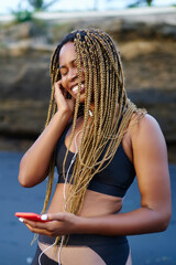 Cheerful afro american young woman happy from audio songs listening in earphones standing on beach during morning run.Dark skinned female dressed in sportive wear enjoying music player on smartphone