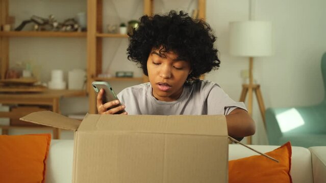 Unhappy angry african woman unpack delivered box receiving damaged shipping mistake problem in postal service. Annoyed confused customer client receiving wrong parcel checking online app on smartphone