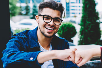 Cheerful hipster guy in eyewear bumping fists in sign of greeting with female friend having meeting...