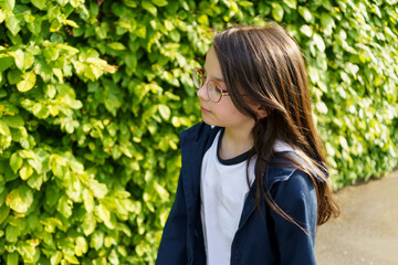 A little beautiful girl in red glasses stands against the background of a green hedge.
