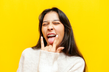 young cheerful asian girl with braces showing rock gesture with hands and tongue on yellow isolated...