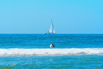 Naklejka premium Surfer Girl Riding Wave Towards Ocean with Sailboat
