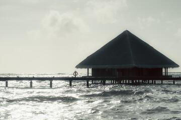 Scenic view of an overwater bungalow with a thatched roof, connected to a wooden pier over the ocean. The sun is reflecting on the water, creating a serene and idyllic atmosphere of a luxury resort