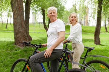 elderly senior couple rides bicycle in the park in the summer and smiles, old gray-haired man and woman are actively resting outdoors, old people practice cycling in forest