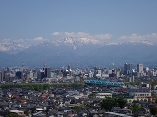 5月新緑の木々と富山市街地の後方に見える残雪の北アルプス薬師岳