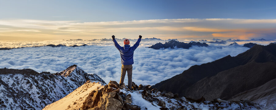 Wide panorama, man on top of a mountain peak, his arms in the air, celebrating success and achievement.