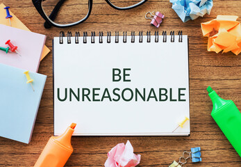 A motivational note stating BE UNREASONABLE displayed on a desk amid colorful stationery items.