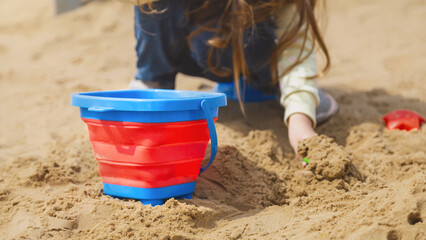 Pretty child girl playing in sand on outdoor playground. Beautiful baby in jeans trousers having fun on sunny warm summer day. Child with colorful sand toys. Healthy active baby outdoors plays games
