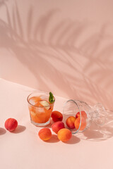 Glass with iced refreshing peach juice drink, peaches and apricots scattered on bright peachy pink...