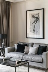 A framed black and white figurative painting is hanging on the wall of an elegant living room. A grey sofa with beige cushions and a glass coffee table are in the room. Light brown curtains.