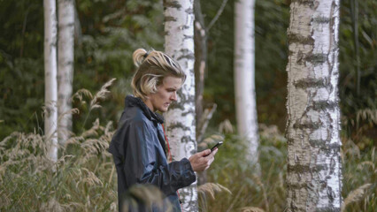 Fototapeta premium Man walks in woods with phone. Stock. Young man is walking along coordinates in phone in forest. Man is looking for way out of forest by phone