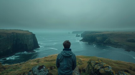 A man gazes at the sea from a rocky coast on a foggy day, with cliffs and distant rock formations visible.