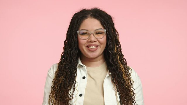 A joyful woman with curly hair and glasses, dressed in a white jacket, is happily blowing kisses and smiling warmly against a pink backdrop. She has a positive and welcoming demeanor