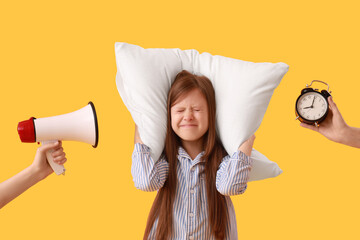 Displeased little girl holding pillow and hands with alarm clock and megaphone on yellow background