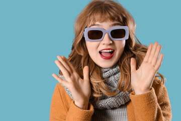 Happy young woman in warm scarf with sunglasses on blue background, closeup