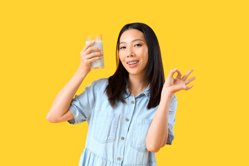 Young Asian woman with glass of milk showing OK on yellow background