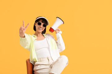 Young woman with megaphone showing victory gesture while sitting on chair against yellow background