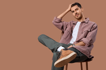 Handsome young man in stylish clothes sitting on stool and posing against beige background