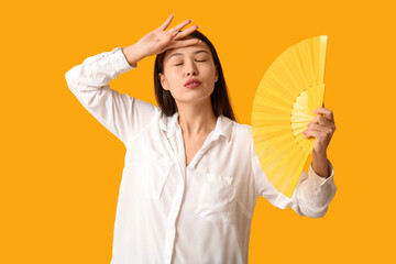 Young woman with hand fan suffering from heat on yellow background