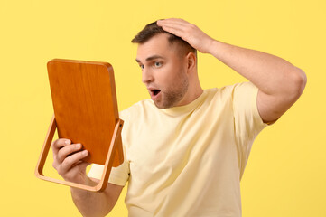 Shocked young man with hair loss problem looking in mirror on yellow background