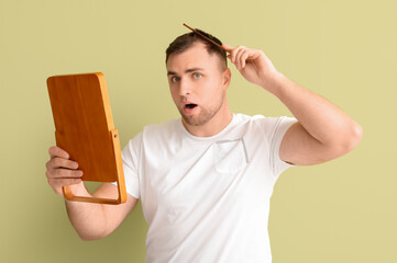 Shocked young man with hair loss problem and mirror combing on green background