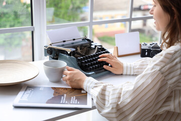 Young woman with coffee cup typing on vintage typewriter at home, closeup