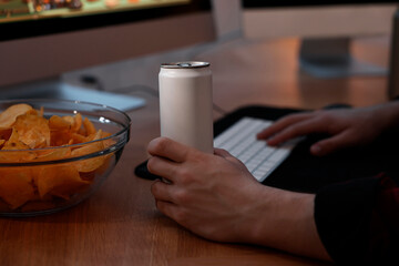 Young man with energy drink and chips playing video game at wooden desk indoors, closeup