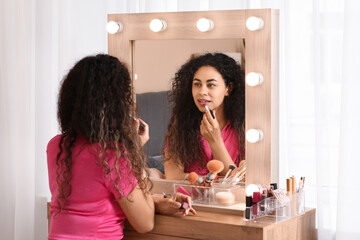 Beautiful young African-American woman with lipstick doing makeup near mirror at home