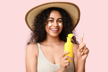 Beautiful young happy African-American woman with bottle of sunscreen cream on pink background