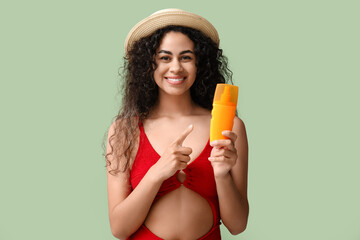 Beautiful young happy African-American woman pointing at bottle of sunscreen cream on green background