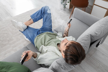 Young man smoking disposable electronic cigarette and chatting with mobile phone on sofa at home