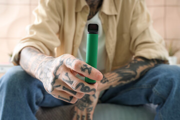 Young tattooed man with disposable electronic cigarette sitting on bed at home, closeup