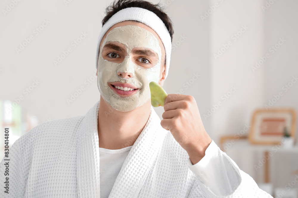 Poster young man with applied clay mask and facial massage tool in bathroom, closeup