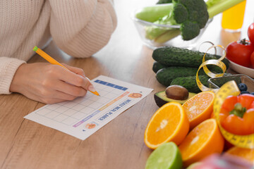 Young woman with healthy food making meal plan on table in kitchen, closeup. Diet concept