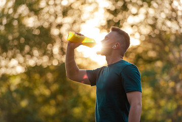 A fit young man drinking water from a bottle after a run or workout in a summer park active healthy...