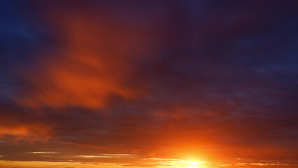 A breathtaking sunrise with intense orange and red tones dominating the sky, transitioning into deeper blues. The sun's rays break through the clouds, creating a warm, glowing effect. 
