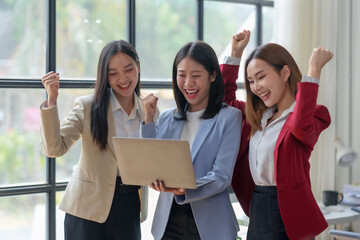 Three cheerful businesswomen in formal attire celebrate success while looking at a laptop in a...