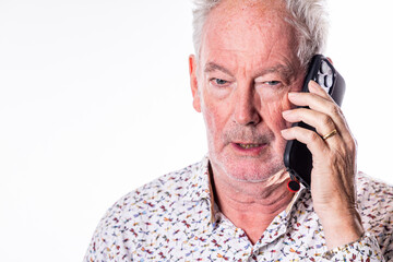 A close-up portrait of an elderly man with white hair and a beard, engaged in a phone conversation. The man is wearing a patterned shirt and holding a smartphone to his ear with a serious and