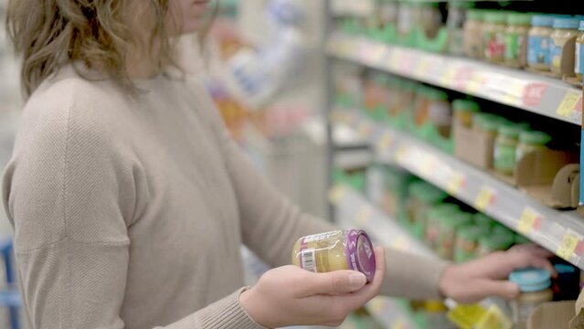 A woman examines jars of baby food in a grocery store aisle, carefully comparing different options. The scene highlights the variety of baby products available for parents.