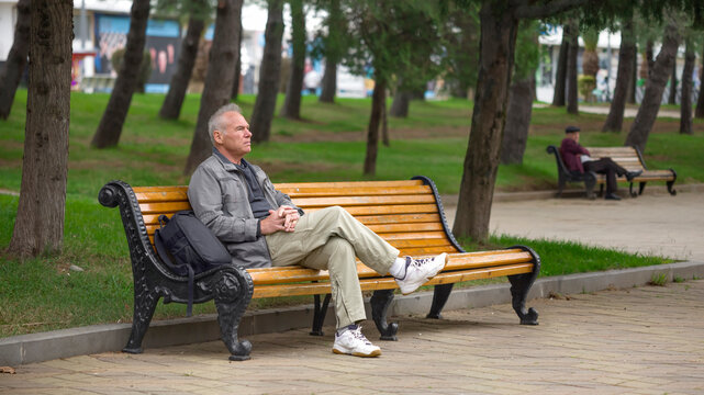 An elderly man with a backpack is sitting on a bench in a city park