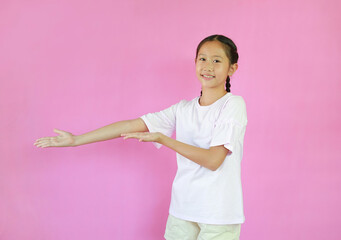 Smiling Asian girl child standing on pink studio background with hand receiving or giving gesture concept. Showing copy space, presenting advertisement.