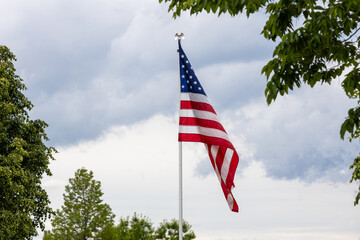 Close up view of an American flag on a flagpole, partially bordered with trees and with overcast sky