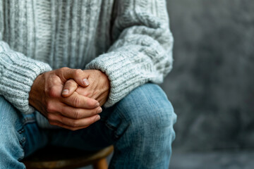 Close up of hands holding a painful knee joint, a man's leg is wounded and shoulders are shown, a closeup of a hand, sitting down against a grey background, wearing a white sweater and blue jeans, spa