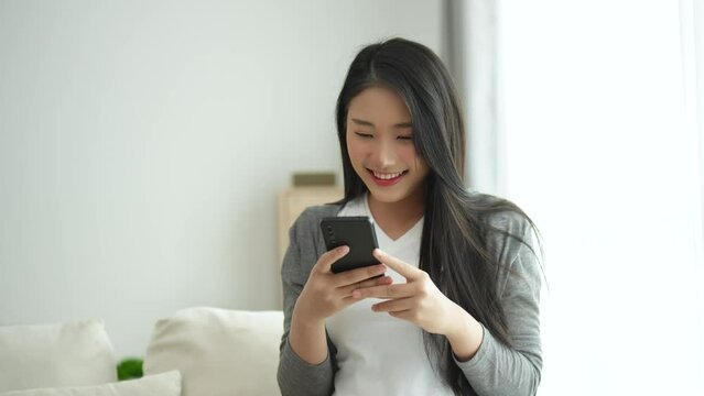 Asian woman hold mobile phone text or message sitting on coach sofa in the living room. Woman smiling use smartphone to browse web, read e-book, trade stocks at home.