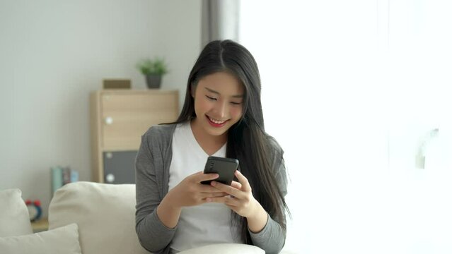 Asian woman hold mobile phone text or message sitting on coach sofa in the living room. Woman smiling use smartphone to browse web, read e-book, trade stocks at home.