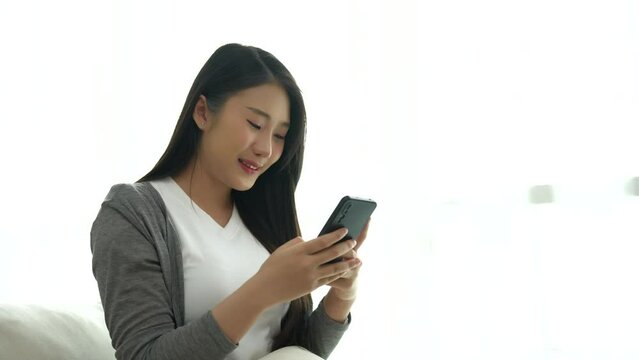 Asian woman hold mobile phone text or message sitting on coach sofa in the living room. Woman smiling use smartphone to browse web, read e-book, trade stocks at home.